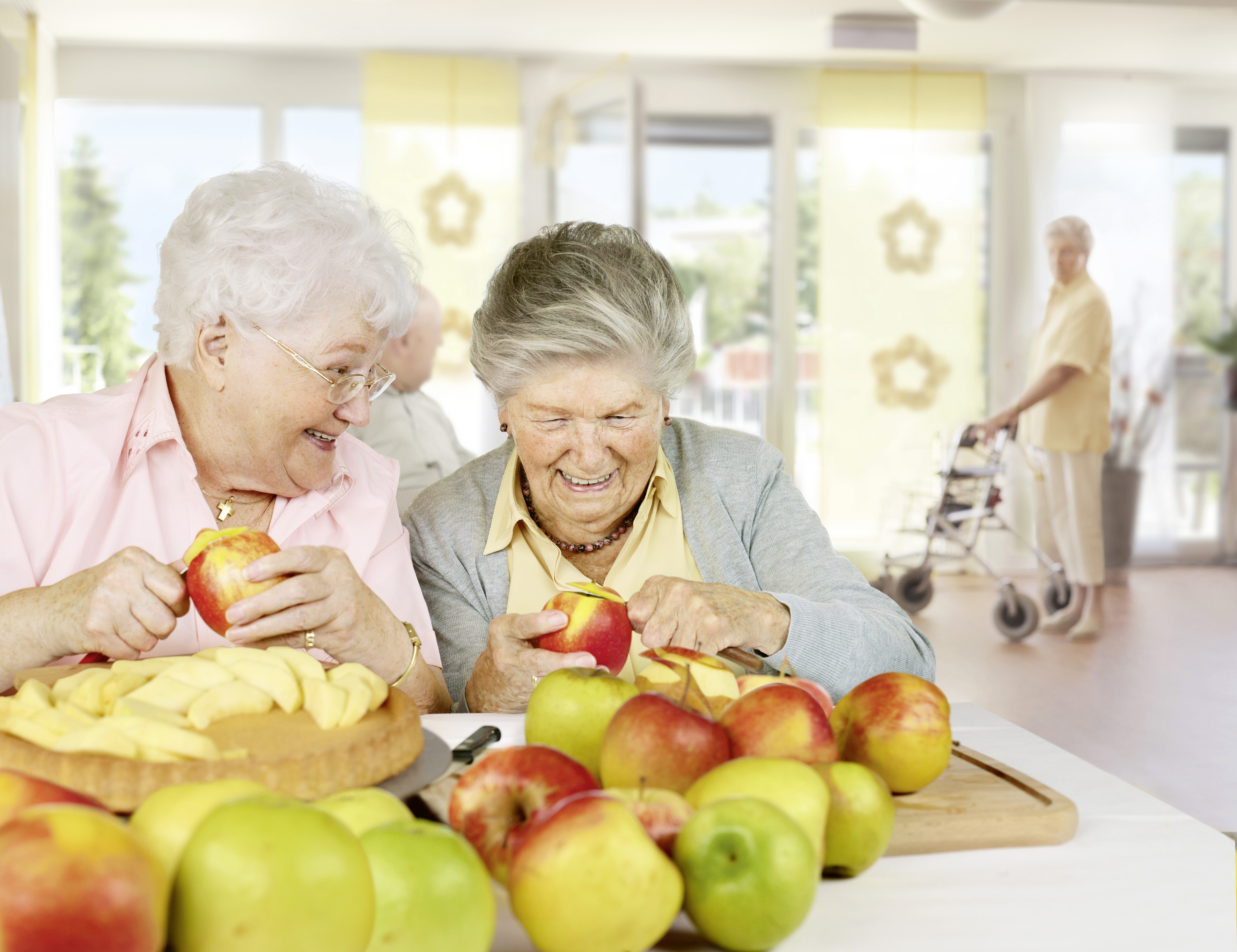 Zwei Seniorinnen in einem Heim schälen Äpfel,um einen Kuchenboden zu belegen.
