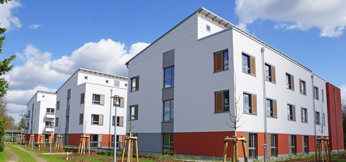 Das Foto zeigt das neue ASB Wohnpflegezentrum Lebensraum für Senioren in Zeuthen. Die Hausfassade ist weiß und rot. Die Anlage ist von jungen Bäumen umgeben.