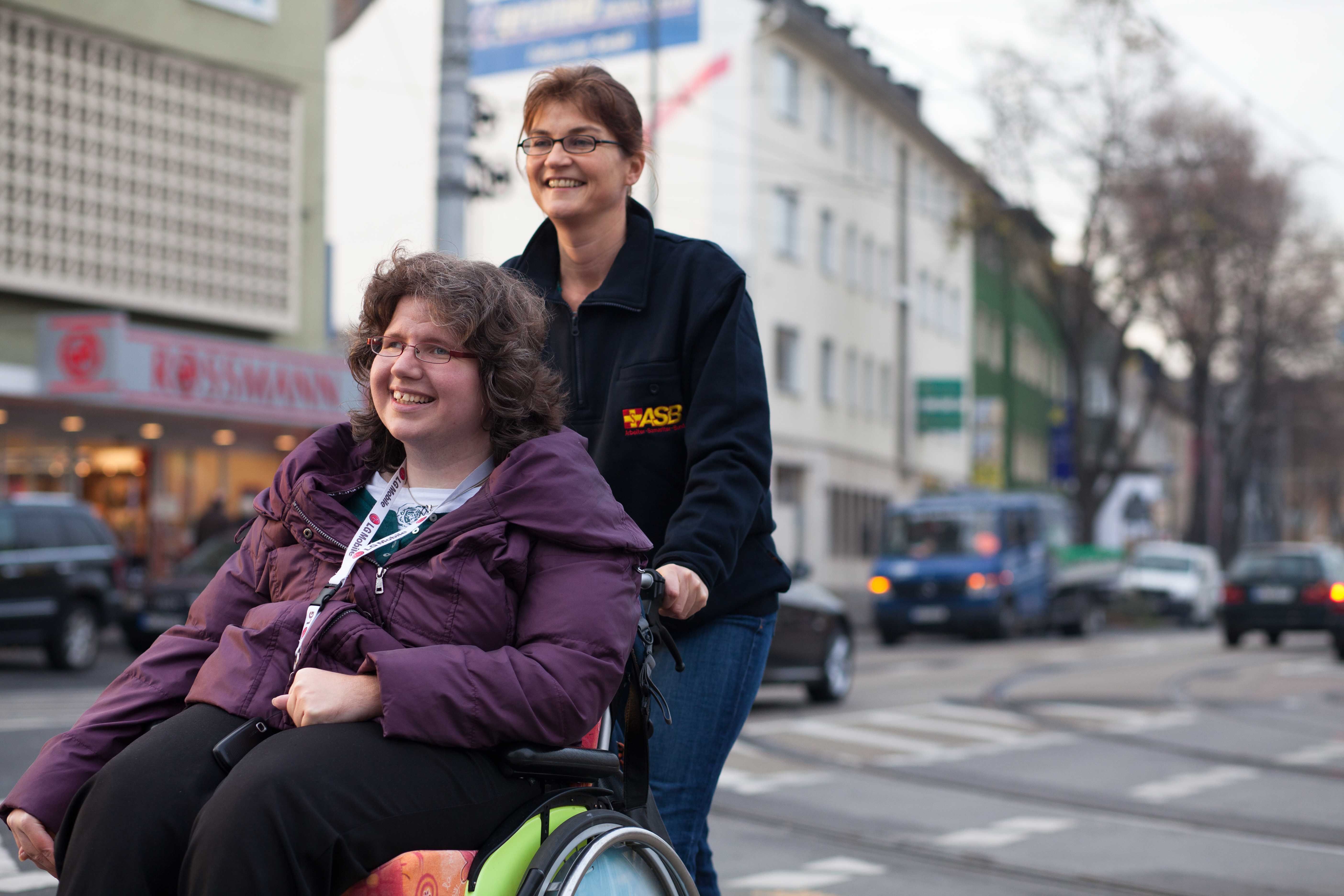 Eine Frau schiebt eine junge Frau im Rollstuhl durch eine Straße.