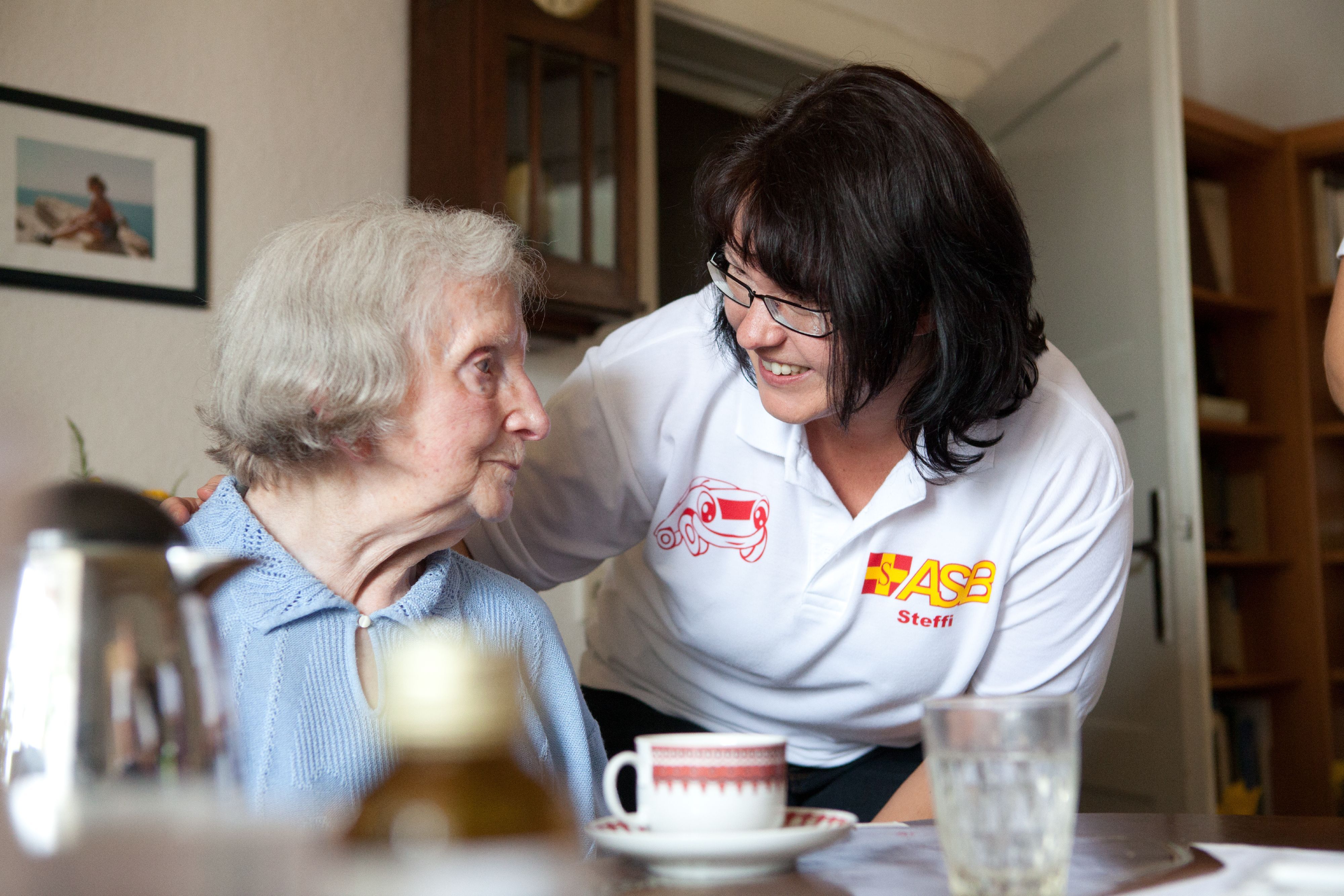 Eine ASB-Pflegerin beugt sich lächelnd zu einer Seniorin,die an ihrem Esstisch Kaffee trinkt.