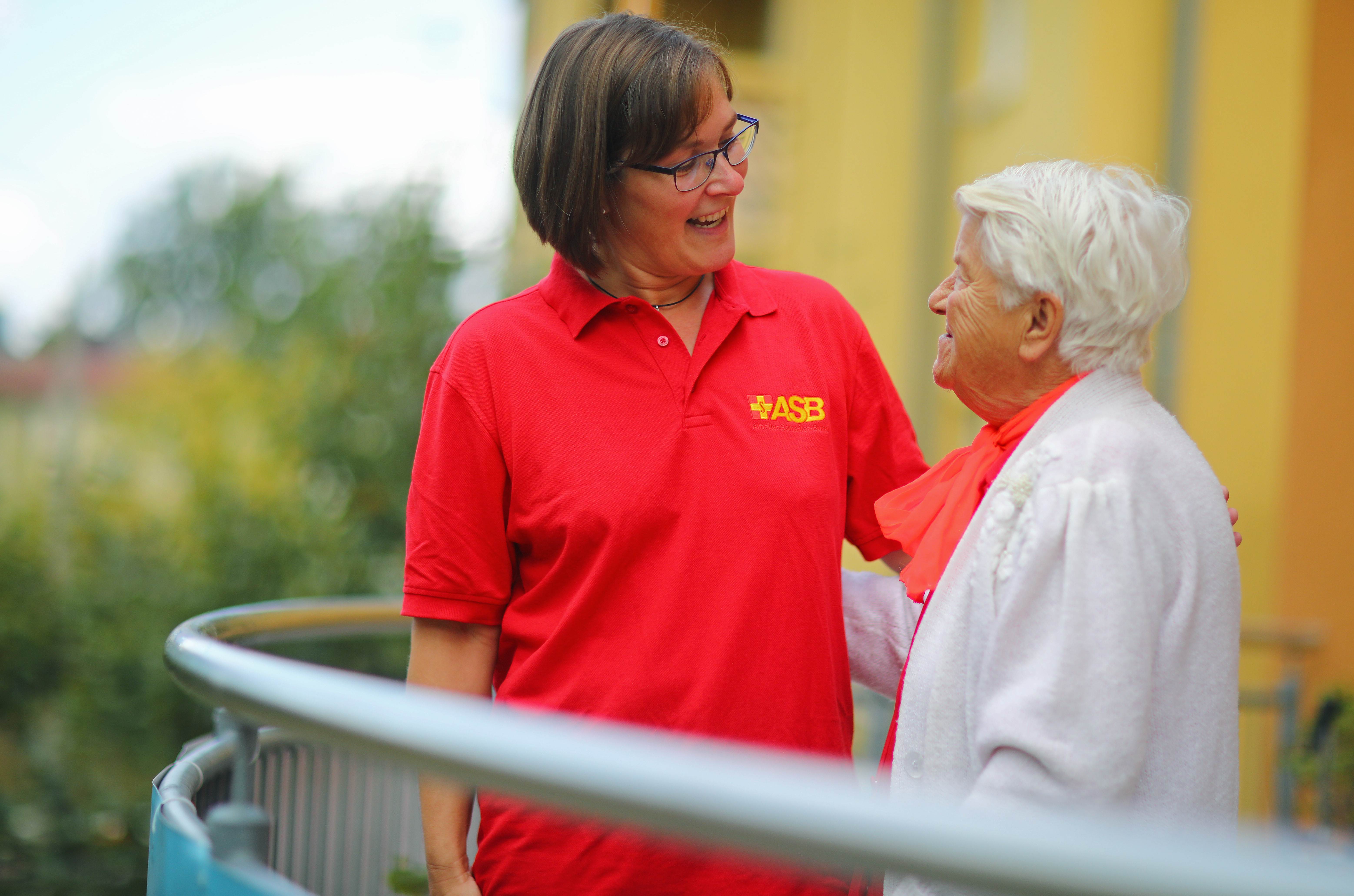 Eine ASB-Pflegerin beugt sich lächelnd zu einer Seniorin,die an ihrem Esstisch Kaffee trinkt.